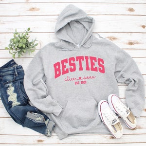 May include: Gray hooded sweatshirt with the text "BESTIES" in red, with the names "alice" and "anna" and the date "EST. 2020" underneath.