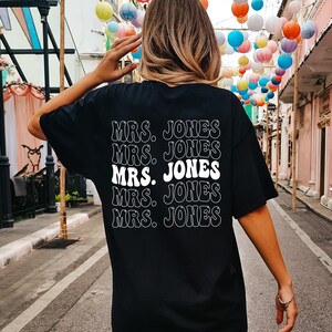 May include: Black t-shirt with the text "MRS. JONES" repeated five times in a stacked design. The text is in a white, retro-style font. The shirt is worn by a person with light brown hair.