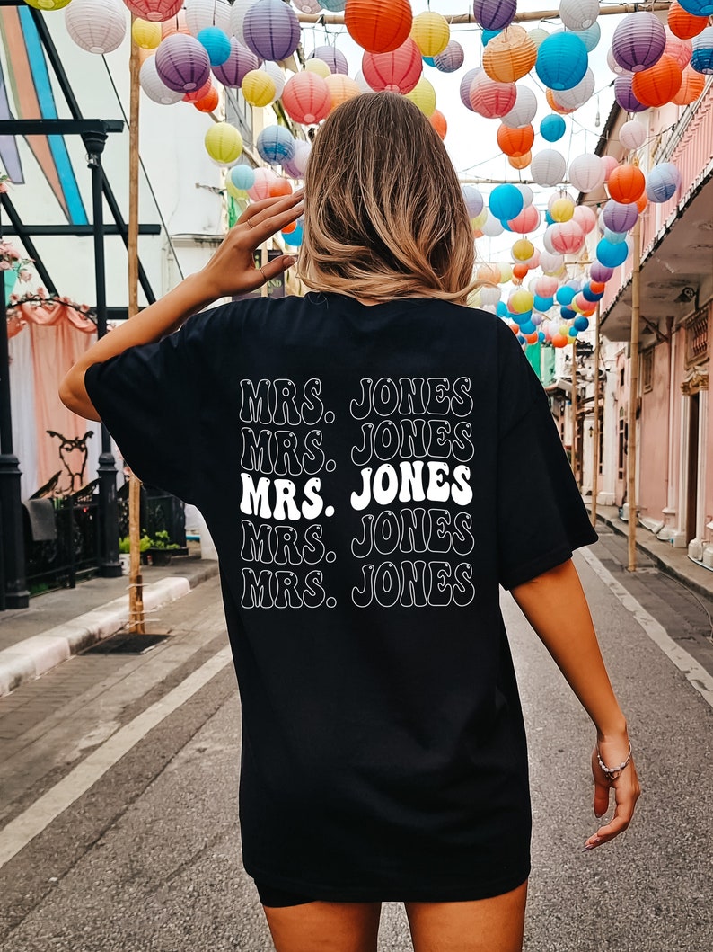 May include: Black t-shirt with the text "MRS. JONES" repeated five times in a stacked design. The text is in a white, retro-style font. The shirt is worn by a person with light brown hair.