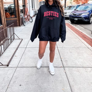 May include: A black hoodie with a red graphic that says "Besties" with a heart and the text "alive & free est. 2020". The person is wearing white sneakers.