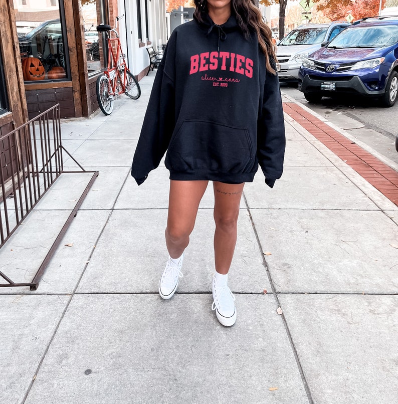 May include: A black hoodie with a red graphic that says "Besties" with a heart and the text "alive & free est. 2020". The person is wearing white sneakers.