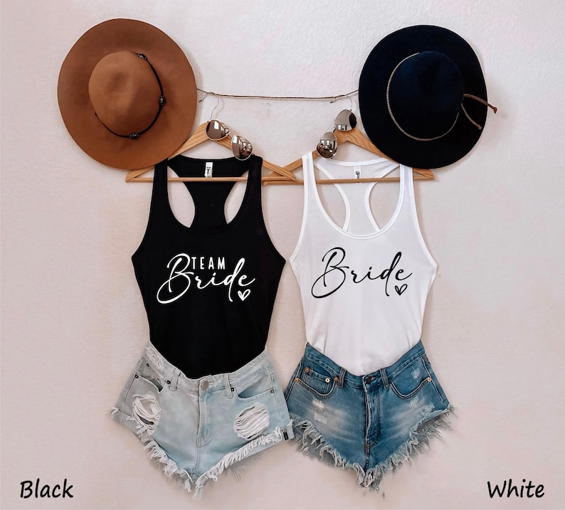 May include: Two tank tops with the text "Team Bride" and "Bride" in white script on a black and white tank top, respectively. The tank tops are hanging on a rack with hats on either side.