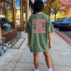 May include: A green oversized t-shirt with the text "Partner in Crime" in pink retro lettering on the back. The shirt is worn by a person with long dark hair and white sneakers.