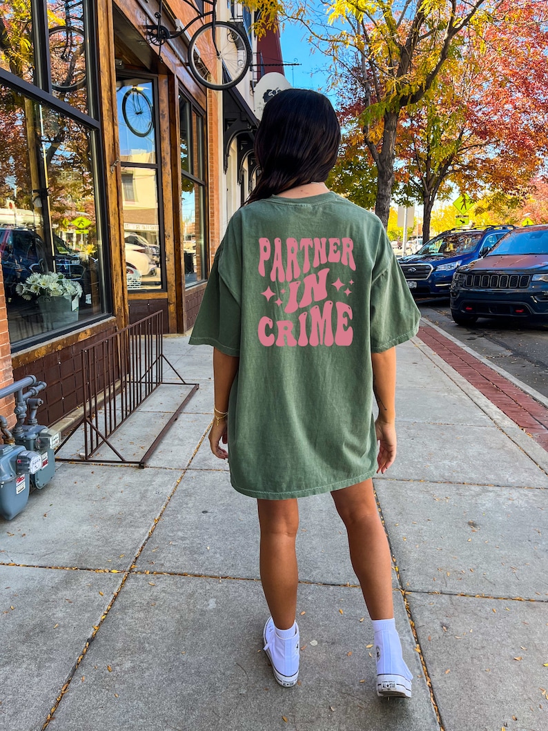 May include: A green oversized t-shirt with the text "Partner in Crime" in pink retro lettering on the back. The shirt is worn by a person with long dark hair and white sneakers.