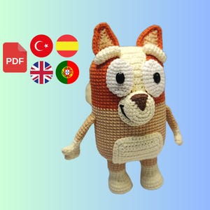 Peut inclure: Peluche en crochet en forme de chien avec une fourrure brune et beige, de grands yeux blancs et un nez brun. Le jouet est debout sur un fond bleu clair.