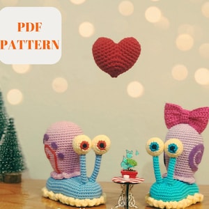 May include: Two crocheted snail amigurumi toys with big eyes, one blue and one purple, sitting on a table with a small potted plant and a red heart floating above them. The text "PDF PATTERN" is in a white box in the upper left corner of the image.