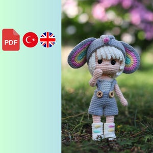 Könnte beinhalten: Eine gehäkelte Puppe mit einem regenbogenfarbenen Hut und einer Latzhose. Die Puppe hat weißes Garnhaar und einen süßen Ausdruck. Die Puppe steht auf einem Bett aus grünem Gras.