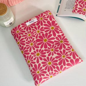 Peut inclure: Une pochette en tissu rectangulaire avec un motif répétitif de fleurs roses ressemblant à des marguerites avec des cœurs jaune moutarde sur fond crème. Une petite étiquette en tissu blanc avec le texte noir Holina est cousue sur le bord.