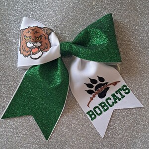 Könnte beinhalten: Grün-weißer Cheer-Bogen mit einem braunen und weißen Bobcat-Maskottchen auf der einen Seite und einem schwarzen Pfotenabdruck mit dem Wort "BOBCATS" auf der anderen Seite.