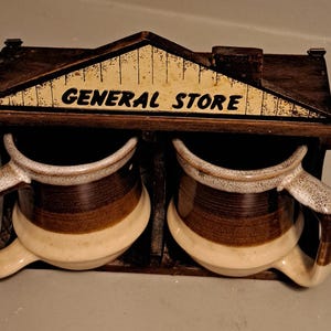 May include: A vintage coffee mug set with two mugs, each with a brown and cream color scheme. The mugs are housed in a wooden holder shaped like a small house, with the words "GENERAL STORE" printed on the front.