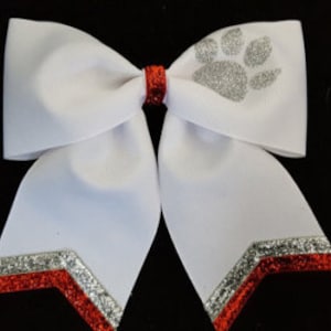May include: White cheer bow with a red and silver glitter chevron design. A silver glitter paw print is centered on the bow.