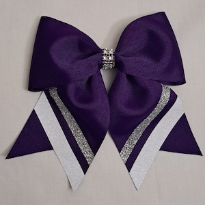 Puede incluir: Un gran lazo de animadora morado con detalles blancos y plateados brillantes. El lazo presenta un centro de diamantes de imitación y un diseño en capas. El lazo está hecho de una tela gruesa y acanalada.