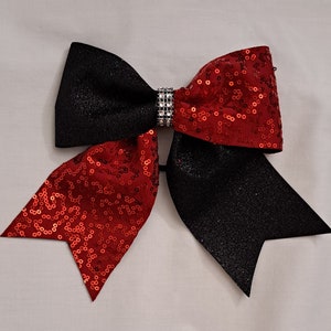 Puede incluir: Un gran lazo de animadora con una combinación de colores negro y rojo. El lazo presenta un material negro brillante en los lados izquierdo y derecho, y lentejuelas rojas en el centro. Una banda de diamantes de imitación plateados está en el centro.