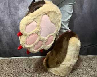 May include: A pair of furry brown and beige paw-shaped boot covers with red claws. The boot covers are worn on a person's legs.