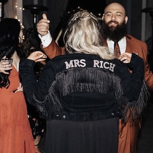 Puede incluir: Una mujer lleva una chaqueta vaquera negra con flecos y las palabras "MRS RICH" en pedrería en la espalda. Está bailando con un hombre que lleva un traje marrón.