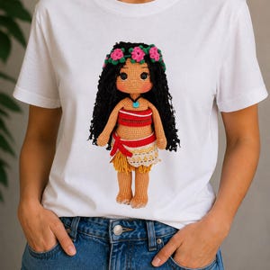 May include: A white short-sleeved t-shirt featuring a graphic print of a crocheted doll with long black hair, a pink flower crown, a red and white patterned top, and a tan skirt with a red waist tie, worn with blue denim jeans.