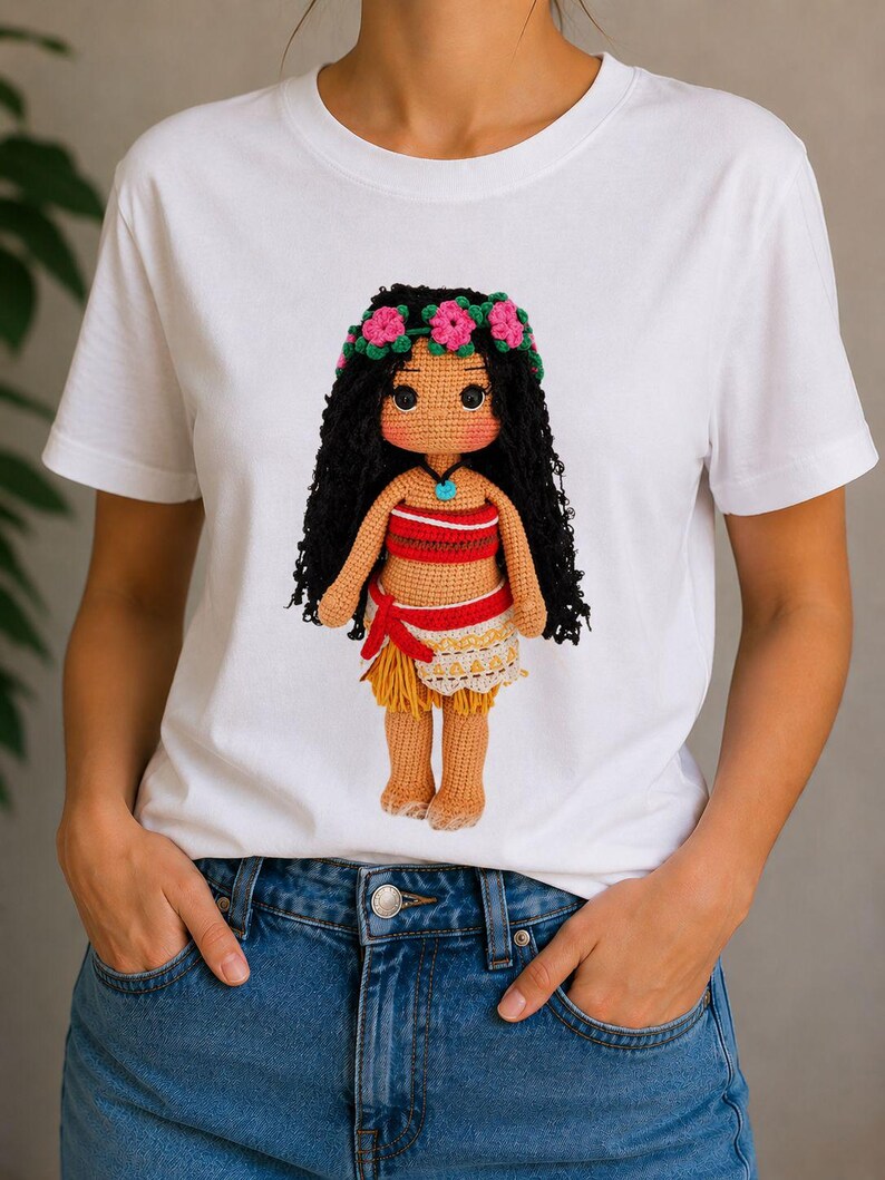 May include: A white short-sleeved t-shirt featuring a graphic print of a crocheted doll with long black hair, a pink flower crown, a red and white patterned top, and a tan skirt with a red waist tie, worn with blue denim jeans.