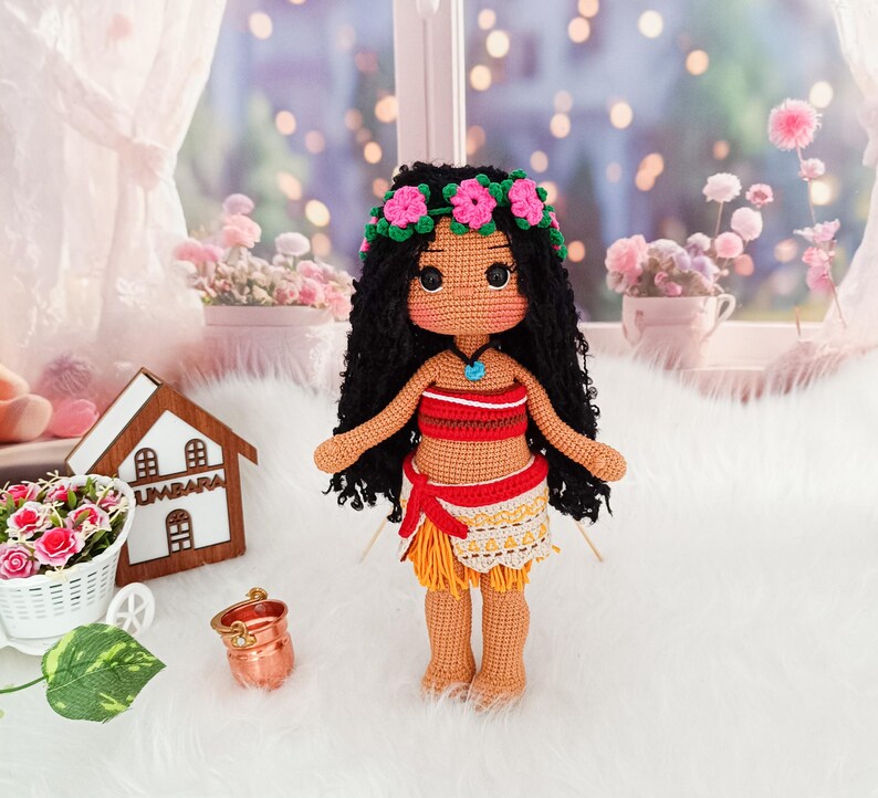May include: Crocheted doll with brown hair, wearing a flower crown, a red and white top, and a yellow skirt with a brown sash. The doll is standing on a white surface.