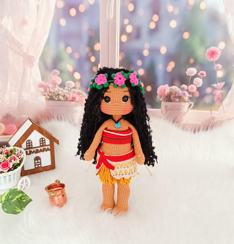 May include: A crocheted doll with brown skin, black curly hair, and a flower crown. She is wearing a red and orange top, a yellow skirt, and a blue necklace. The doll is standing on a white fluffy surface.
