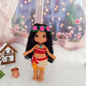 May include: Crocheted doll with brown skin, black curly hair, and a flower crown. She is wearing a red and yellow outfit with a blue necklace. The doll is standing on a white fluffy surface.