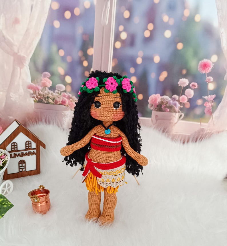 May include: Crocheted doll with brown skin, black curly hair, and a flower crown. She is wearing a red and yellow outfit with a blue necklace. The doll is standing on a white fluffy surface.