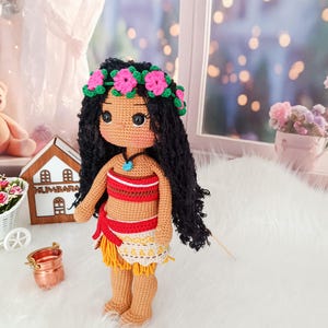 May include: A crocheted doll with brown skin, black curly hair, and a flower crown. She is wearing a red and yellow outfit with a blue necklace. The doll is standing on a white fluffy surface.