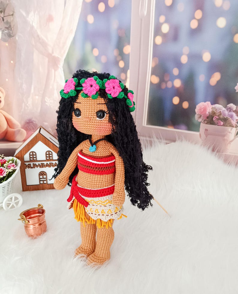 May include: A crocheted doll with brown skin, black curly hair, and a flower crown. She is wearing a red and yellow outfit with a blue necklace. The doll is standing on a white fluffy surface.