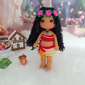 May include: A crocheted doll with long black hair, wearing a red and white top, a yellow skirt, and a flower crown. The doll has brown skin and large brown eyes.