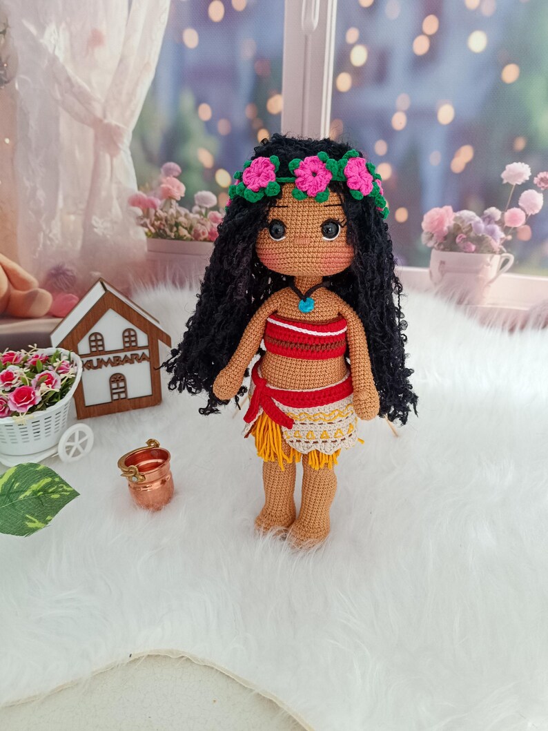May include: A crocheted doll with long black hair, wearing a red and white top, a yellow skirt, and a flower crown. The doll has brown skin and large brown eyes.