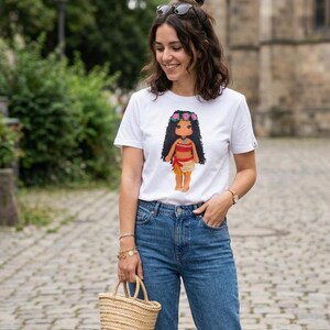 May include: A white short-sleeved t-shirt featuring a graphic print of a doll with long black hair, a flower crown, a red top, and a tan skirt. The shirt is paired with blue denim jeans and a small woven straw handbag with two handles.