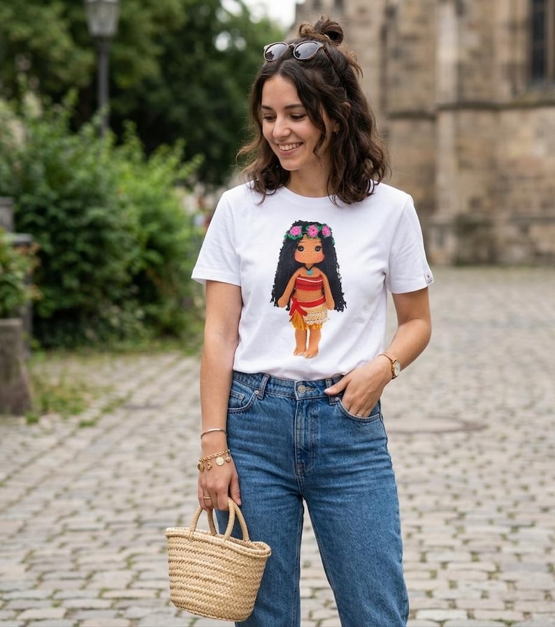 May include: A white short-sleeved t-shirt featuring a graphic print of a doll with long black hair, a flower crown, a red top, and a tan skirt. The shirt is paired with blue denim jeans and a small woven straw handbag with two handles.