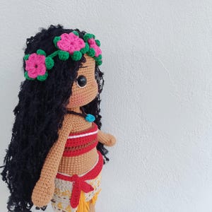 May include: A crocheted doll with brown skin, black curly hair, and a flower crown. The doll is wearing a red and white top and a brown and yellow skirt.