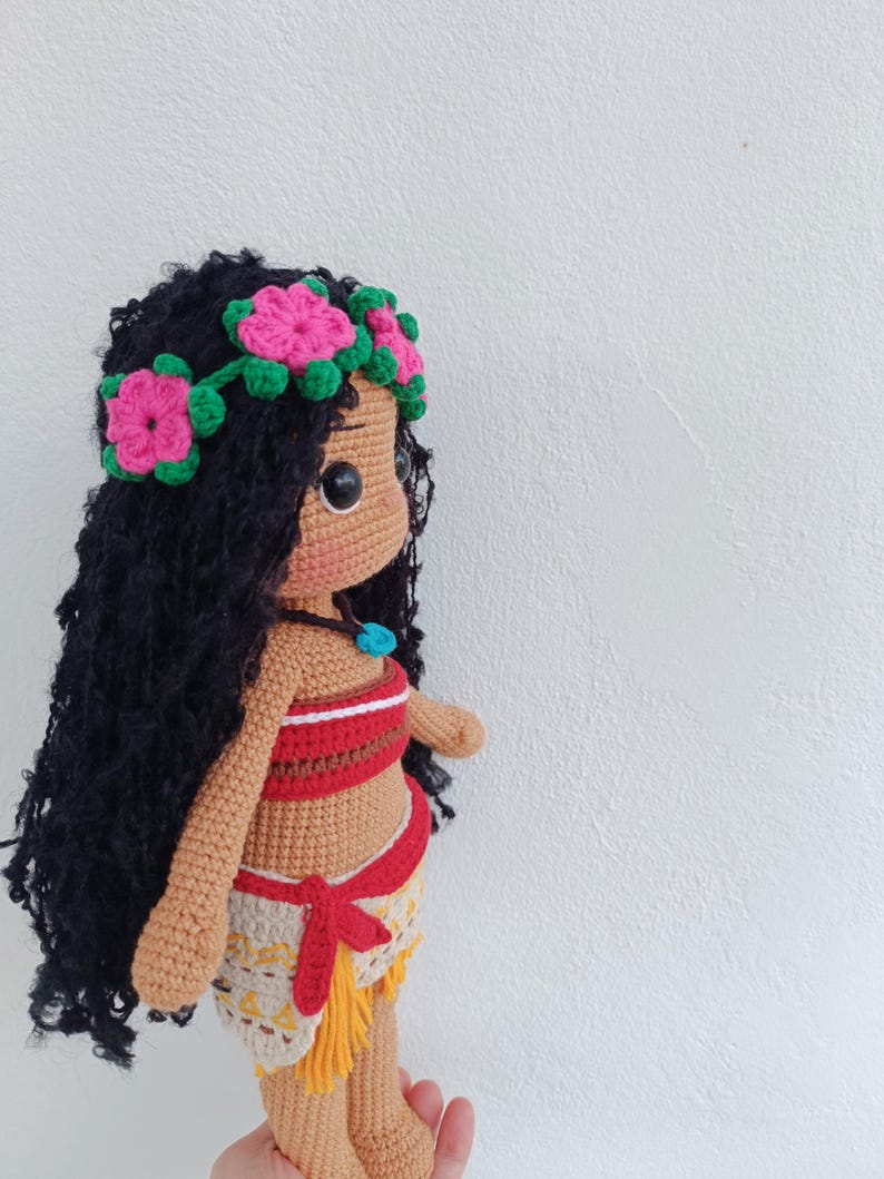 May include: A crocheted doll with brown skin, black curly hair, and a flower crown. The doll is wearing a red and white top and a brown and yellow skirt.