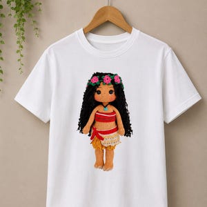 May include: A white short-sleeved t-shirt featuring a centered graphic of a crocheted doll with long black hair, a pink flower crown, a red and tan top, and a tan skirt. The doll has a light brown complexion and a small blue pendant necklace.