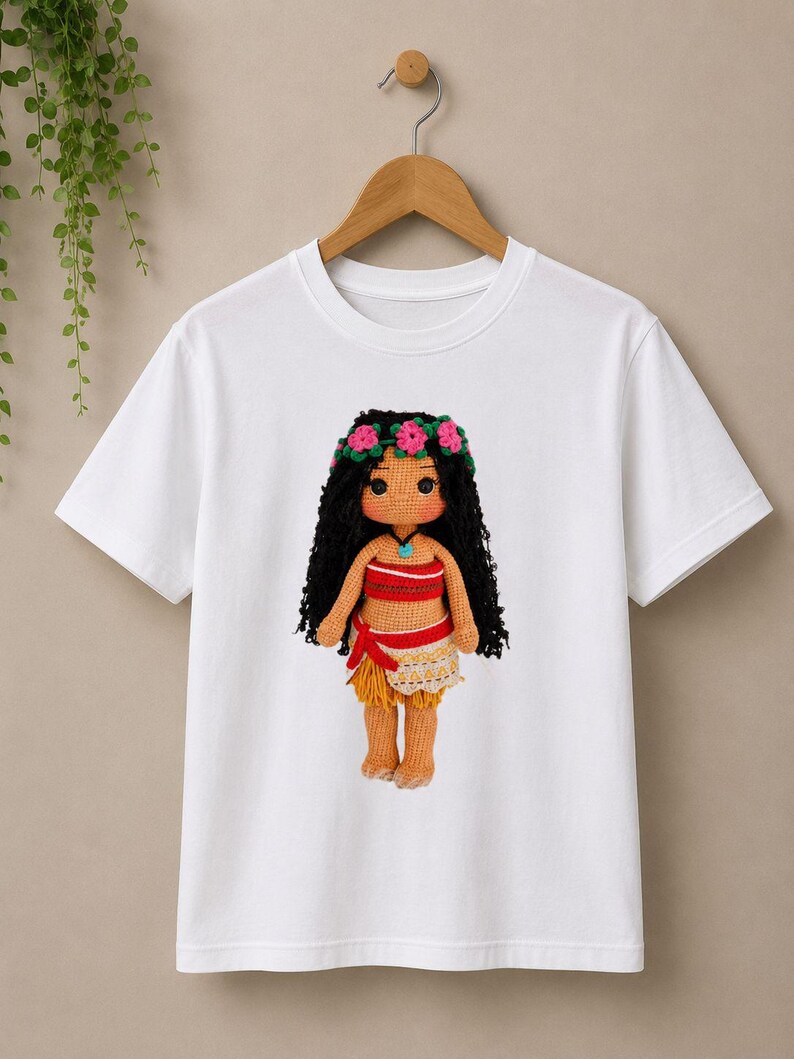 May include: A white short-sleeved t-shirt featuring a centered graphic of a crocheted doll with long black hair, a pink flower crown, a red and tan top, and a tan skirt. The doll has a light brown complexion and a small blue pendant necklace.