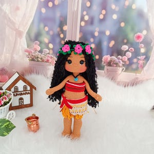 May include: A crocheted doll with brown skin, black curly hair, and a flower crown. She is wearing a red and yellow outfit with a blue necklace. The doll is standing on a white fluffy surface.