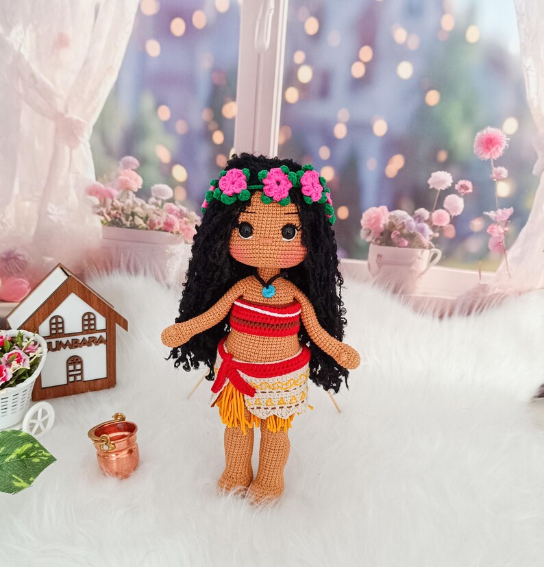 May include: A crocheted doll with brown skin, black curly hair, and a flower crown. She is wearing a red and yellow outfit with a blue necklace. The doll is standing on a white fluffy surface.
