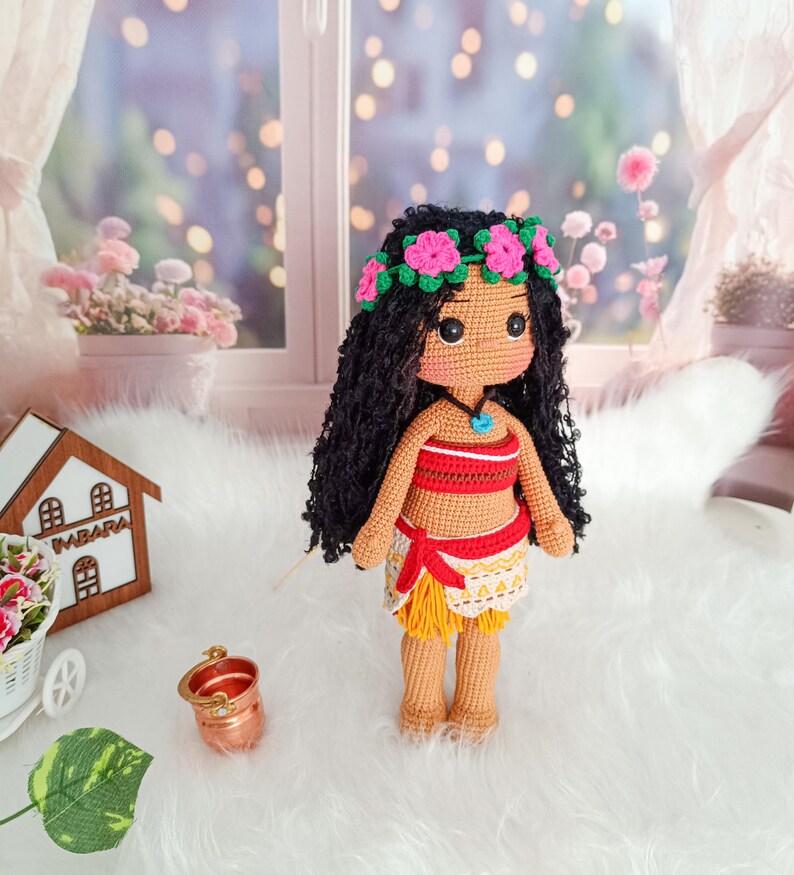 May include: A crocheted doll with brown skin, black hair, and a flower crown. She is wearing a red and white top, a yellow skirt, and a blue necklace.