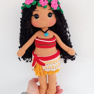 May include: Crocheted doll with brown skin, black hair, and a flower crown. She is wearing a red and white top, a yellow and brown skirt, and a blue necklace.