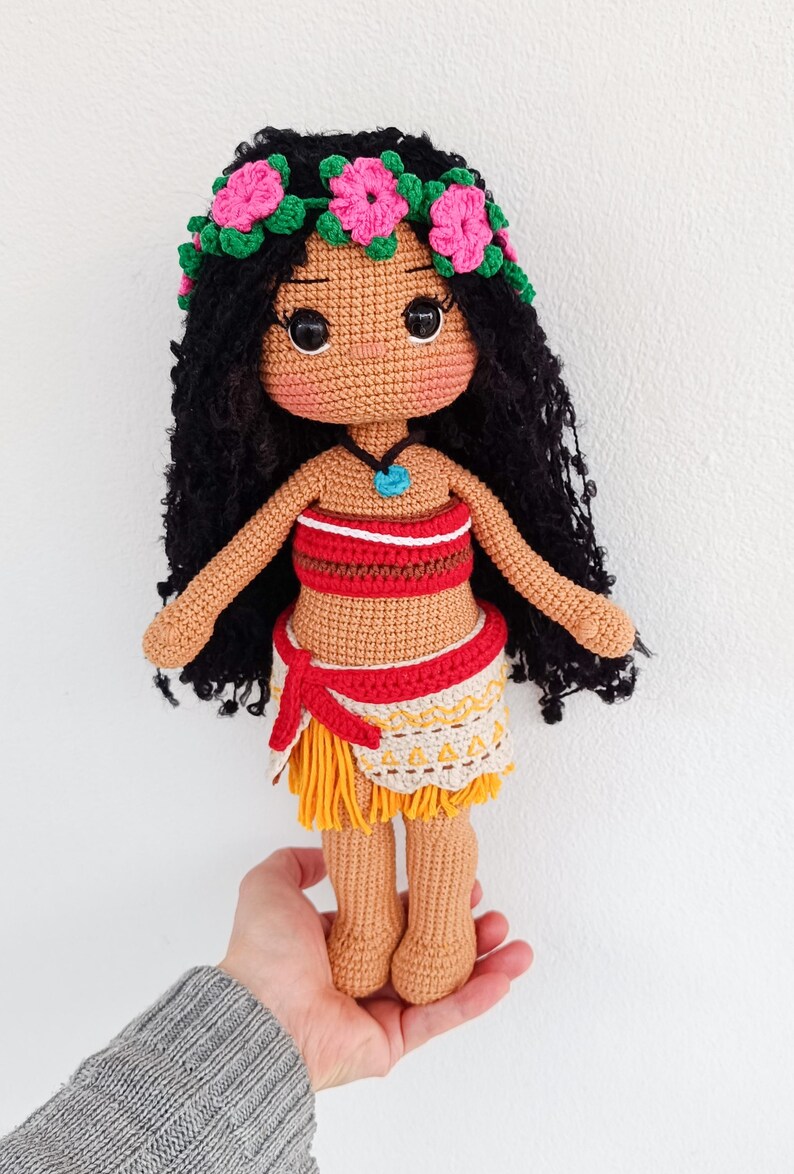 May include: Crocheted doll with brown skin, black hair, and a flower crown. She is wearing a red and white top, a yellow and brown skirt, and a blue necklace.