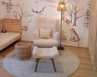 May include: A light wood cot with a white mattress and a white and cream armchair with a white cushion. The wall behind the cot and armchair has a woodland mural with a deer, owl, and other animals. The room has a light wood floor and a light wood cabinet.