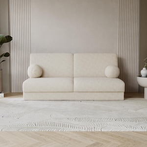 Pode incluir: Um sofá creme de dois lugares com duas almofadas redondas. O sofá está sobre um tapete estampado, com uma grande planta em vaso à esquerda e uma pequena mesa lateral com decoração à direita. As paredes são de cor neutra.