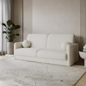 Könnte beinhalten: Ein cremefarbenes Zweisitzer-Sofa mit abgerundeten Armlehnen und zwei passenden runden Wurfkissen. Das Sofa steht auf einem gemusterten Teppich in einer neutralen Farbpalette, mit einer großen Topfpflanze und einem kleinen Beistelltisch in der Nähe.