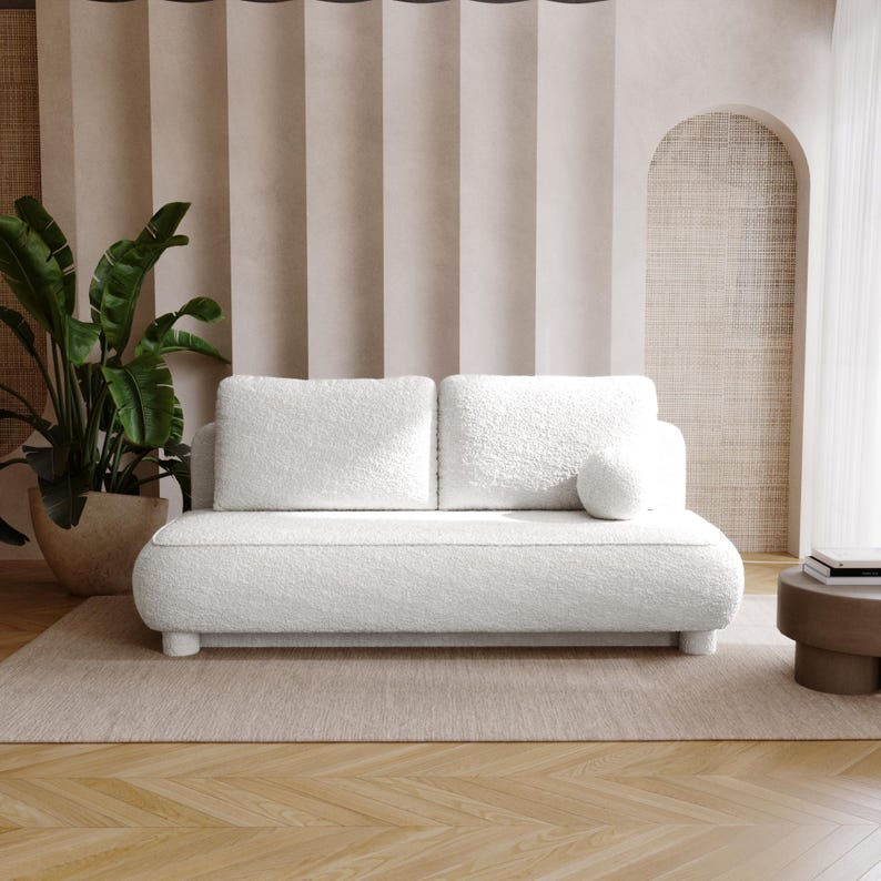 May include: A white, two-seater sofa with a rounded design and a fluffy, textured fabric. The sofa has a matching round bolster pillow and sits on short, cylindrical legs. A large potted plant and a small side table are also visible.