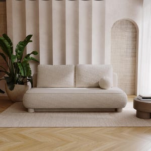 Könnte beinhalten: Ein cremefarbenes Zweisitzer-Sofa mit abgerundeten Kanten und einem passenden runden Zierkissen. Das Sofa steht auf einem hellbeigen Teppich, mit einer großen Topfpflanze links und einem kleinen Beistelltisch rechts. Der Hintergrund zeigt neutrale Vorhänge und eine gewölbte Wand.