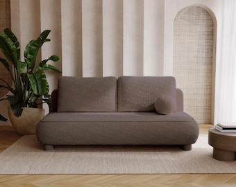 Op de afbeelding: Een moderne, taupekleurige bank met een afgerond ontwerp en een bijpassend rond kussen. De bank staat op een lichtbeige tapijt, met een potplant en een kleine bijzettafel op de achtergrond. De kamer heeft een neutraal kleurenpalet.
