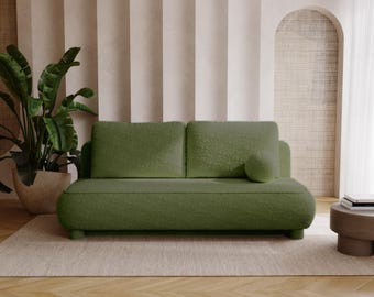 Op de afbeelding: Een groene tweezitsbank met een afgerond ontwerp en een bijpassend rond kussen. De bank staat op een beige tapijt, met een grote plant in pot aan de linkerkant en een kleine bijzettafel aan de rechterkant. De achtergrond heeft neutrale gordijnen en een gewelfde muur.
