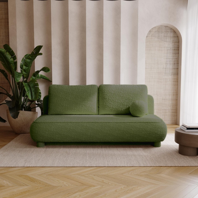 May include: A green, two-seater sofa with a rounded design and a matching round pillow. The sofa is placed on a beige rug, with a large potted plant to the left and a small side table to the right. The background features neutral-toned curtains and an arched wall.