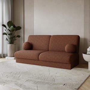 Könnte beinhalten: Ein zweisitziges, rostfarbenes Sofa mit zwei runden Kissen. Das Sofa steht auf einem hellen Teppich mit Wellenmuster. Ein Blumentopf und ein kleiner Beistelltisch sind im Hintergrund zu sehen. Der Raum hat eine neutrale Farbpalette.