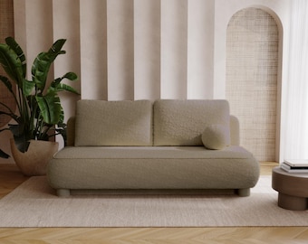 Op de afbeelding: Een moderne, lichtgekleurde bank met een afgerond ontwerp en een textuurrijke, zachte stof. De bank heeft twee rugkussens en een klein, rond sierkussen. De kamer heeft een grote plant in een pot, een kleine bijzettafel en een neutraal getint tapijt.